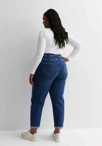 Jeans de tiro alto de color azul oscuro con puños enrollados, diseño de cinco bolsillos y una textura de denim suave. Llevados con una blusa blanca de manga larga y zapatillas.
