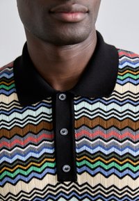 Polo tričko vyrobené z pletenej látky, s viacfarebným zigzag vzorom, čiernym golierom a trojicou zapínacích gombíkov s tmavými gombíkmi.