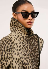 Płaszcz w panterkę z wysokim kołnierzem, w beżowo-czarnych kolorach, o miękkiej teksturze, w zestawieniu z czarnymi kanciastymi okularami przeciwsłonecznymi, które odbijają światło.