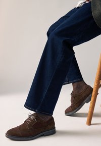 Chaussures habillées en daim marron avec design à bout golf, associées à un jean foncé et des chaussettes texturées. Les chaussures présentent des lacets et une semelle noire contrastante.