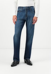 Persona de pie con jeans rectos de color azul oscuro, una blusa blanca de manga larga y zapatos negros de punta frente a un fondo blanco liso.