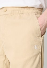Pantalon beige avec taille élastique et logo blanc "cK" près de la poche droite, main insérée dans la poche, porté avec un haut blanc.