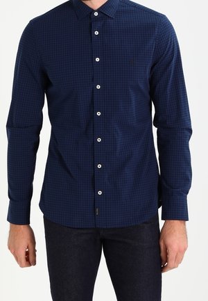 Uomo che indossa una camicia a scacchi blu navy aderente con maniche lunghe e pantaloni scuri su uno sfondo neutro.