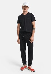 Sort V-hals t-shirt, sorte sweatpants med snøre, og røde sneakers med beige detaljer. Hvid kasket med tekst på forsiden.