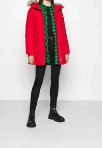 Parka de invierno acolchada roja con capucha forrada de piel, cierre de cremallera y bolsillos, llevada sobre una camisa verde a cuadros y pantalones negros ajustados con botas.