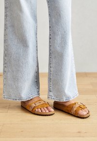 Bruine suède slidersandalen met twee verstelbare banden, kurken voetbed en gestructureerde zolen, gecombineerd met lichtblauwe wijde jeans.