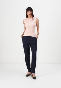 Světlá růžová bezramínková blůza s draped krčkem, spárovaná s tmavě námořnickými kalhotami na míru. Modelka stojí s rukama v kapsách a má na sobě černé podpatky.