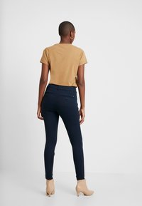 Mörkblå byxor med en åtsittande design, två bakfickor och subtila sömdetaljer, i kombination med en ljusbrun t-shirt med kort ärm.