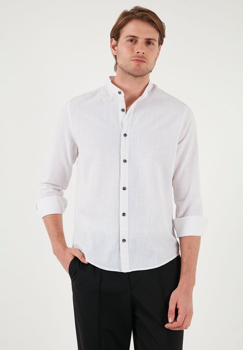Camisa de lino blanca con cuello mao, cinco botones oscuros, mangas largas dobladas y un acabado texturizado. Combinada con pantalones negros.