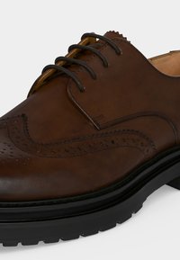Καφέ δερμάτινο παπούτσι brogue με διακοσμητικές τρύπες, μαύρα στρογγυλά κορδόνια και παχύ μαύρο καουτσούκ σόλα.