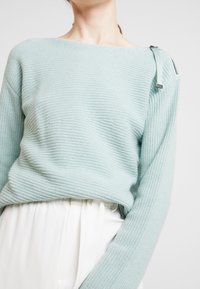Hellblau strukturierter Strickpullover mit weitem Ausschnitt und langen Ärmeln, der ein einzigartiges Riemen-Detail an der Schulter aufweist.