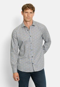 Mann mit kurzen, lockigen Haaren, der ein langärmliges, geknöpftes Hemd mit blauen, orangefarbenen und weißen Wirbelmustern sowie dunkelblauen Jeans trägt.