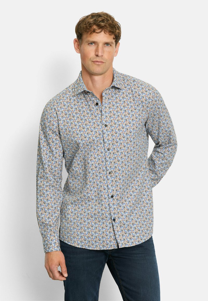 Mann mit kurzen, lockigen Haaren, der ein langärmliges, geknöpftes Hemd mit blauen, orangefarbenen und weißen Wirbelmustern sowie dunkelblauen Jeans trägt.