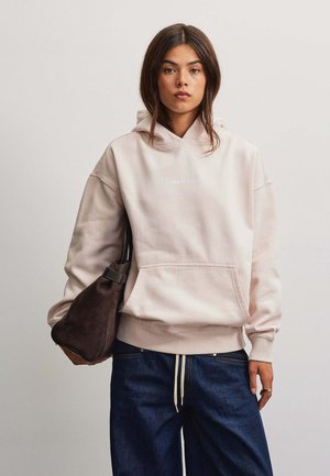 Junge Frau mit langen dunklen Haaren trägt einen hellbeigen Hoodie und dunkelblaue Jeans und hält eine große dunkelbraune Schultertasche am linken Arm.