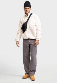Weiße Fleece-Jacke mit Reißverschluss, graue weite Hose, braune Schuhe und schwarze Mütze. Schwarze Umhängetasche mit strukturiertem Muster.
