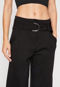 Svarta vida byxor med bälte i midjan som har en silverfärgad D-ring, sidofickor och en mjuk tygstruktur.
