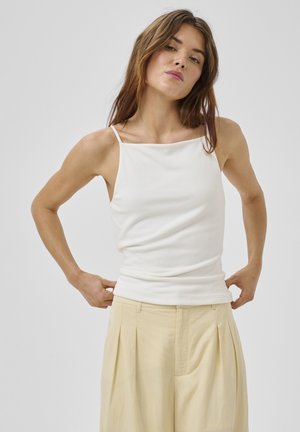 Młoda kobieta poprawiająca biały top bez rękawów, nosząca beżowe plisowane spodnie z wysokim stanem, stoi na tle jednolitego jasnego tła.