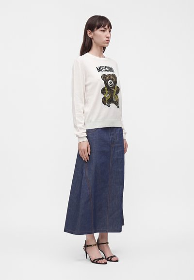 Femme portant un sweat-shirt blanc Moschino avec un motif d'ours en peluche, une longue jupe en jean et des sandales à talons hauts noires, debout sur fond blanc.