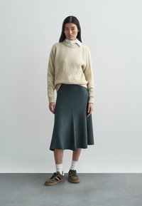 Jasnobeżowy dziany sweter, ciemnozielona rozkloszowana spódnica, biała koszula z kołnierzykiem pod spodem oraz oliwkowo-zielone trampki z białymi i granatowymi paskami.