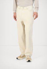 Beige džinsi, brīvs piegriezums, ar augstu jostasvietu, piecu kabatu dizainu un metālisku pogas aizdari. Valkāti ar beigu sporta apaviem.