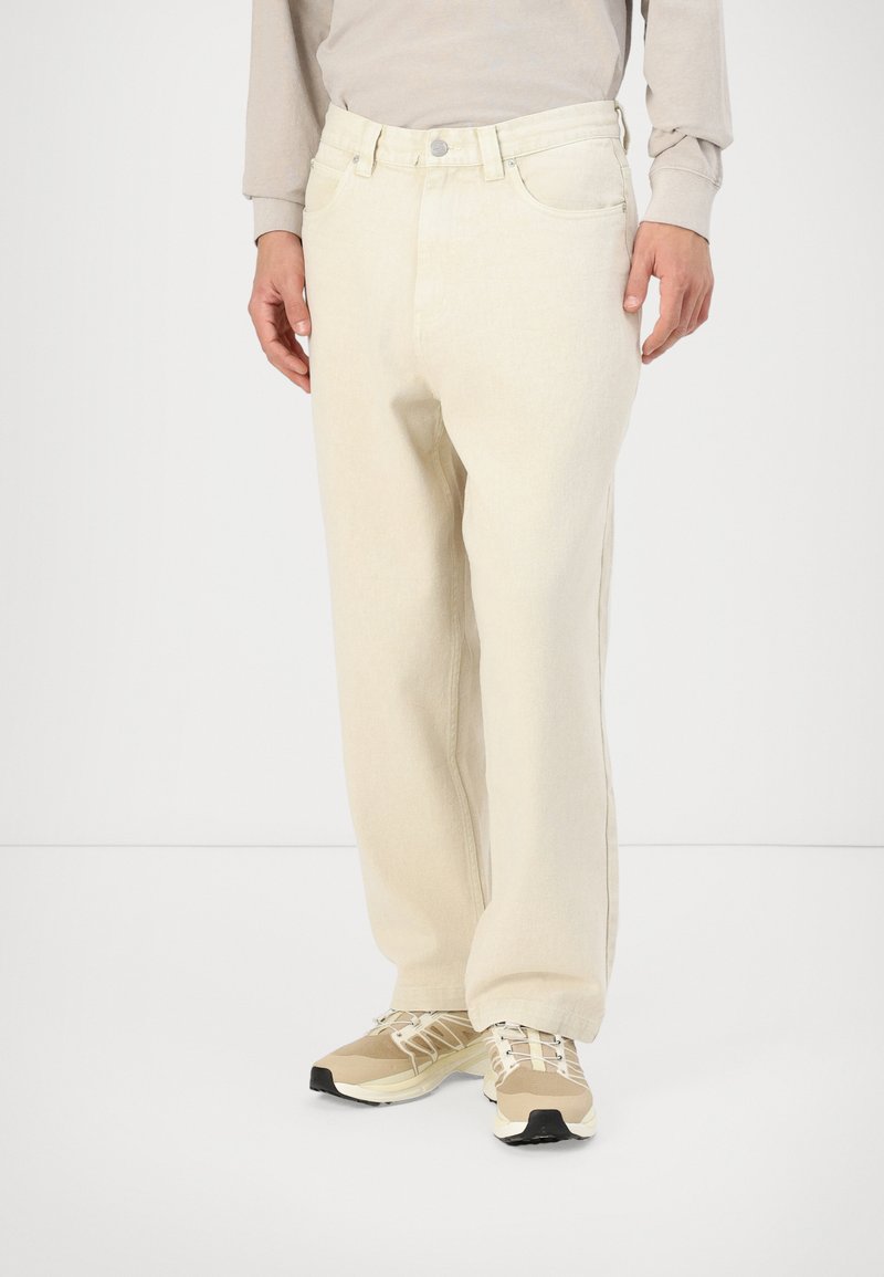 Beige džinsi, brīvs piegriezums, ar augstu jostasvietu, piecu kabatu dizainu un metālisku pogas aizdari. Valkāti ar beigu sporta apaviem.