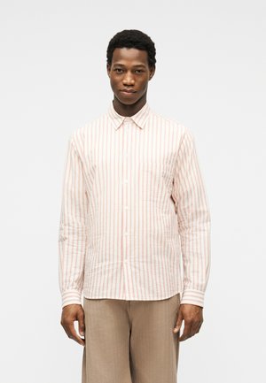 Moški v beli dolgi srajci z gumbi, s tankimi rdečimi navpičnimi črtami in bež hlačah, ki stoji pred enobarvnim ozadjem.