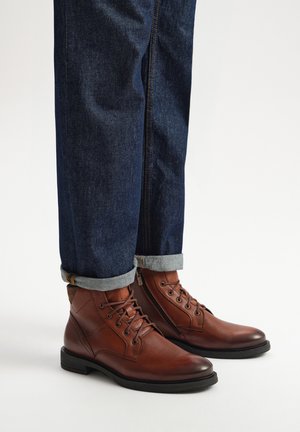 Brązowe skórzane botki za kostkę z zaokrąglonym noskiem, sznurowanym designem i czarną gumową podeszwą, zestawione z podwiniętymi ciemnymi dżinsami.