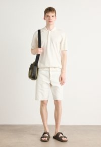 Polo beige, short en denim blanc, sandales à glissière marron et sac bandoulière vert olive. Design minimaliste, textures douces.