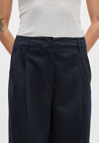 Egyedi szabású sötét tengerészkék nadrág magas derékkal, pliszírozott elülső résszel és oldalzsebekkel. Sima szövetből készült, tiszta kidolgozással.