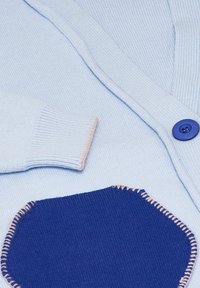 Cardigan azzurro chiaro con texture a costine, dotato di una tasca blu con cuciture rosa e un bottone blu rotondo sul davanti.