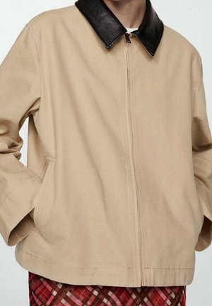 Veste beige zippée avec col en cuir noir portée sur une chemise blanche, associée à une jupe ou un pantalon à carreaux rouge et marron.