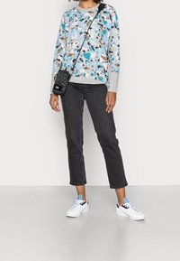 Personne portant une chemise à manches longues à motifs multicolores, un jean noir court, des baskets blanches avec des accents bleus, et un petit sac bandoulière noir.