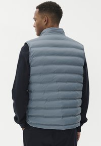 Vīrietis stāv sānis, ģērbies gaiši zilā nošūtā bezpalīdzīgā veste pāri tumšai garai piedurkņu džemperim pret vienkāršu fona.