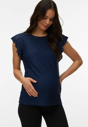 Zwangere vrouw in een marineblauw topje met oogjeskant en zwierige mouwen, met één hand haar buik vasthoudend, staand tegen een effen witte achtergrond.