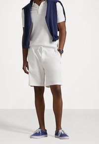 Shorts en coton blanc avec cordon de serrage, associés à un polo blanc et un pull marine porté sur les épaules. Chaussures : baskets en toile bleue.