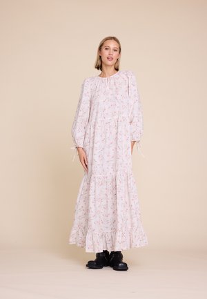 Kvinne iført en lang, lys rosa blomsterkjole med puffermer og svarte snøresko, stående mot en beige bakgrunn.