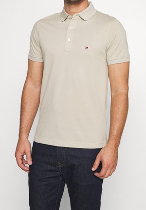 Homme portant un polo beige à manches courtes avec des boutons et un petit logo sur la poitrine, associé à un jean bleu foncé.