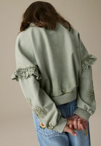 Grüner Sweatshirt mit gerafftem Spitzenbesatz an den Schultern und floraler Stickerei an den Ärmeln, kombiniert mit hellblauen Jeans.