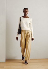 Top cu dungi deschise, cu mâneci lungi, pantaloni bej plisați și sandale negre cu talpă joasă. Design simplu, cu o croială lejeră și texturi moi.