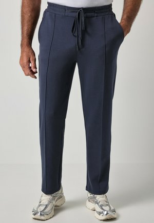 Uomo che indossa pantaloni blu navy con coulisse e sneakers bianche, in piedi con le mani in tasca su uno sfondo chiaro e uniforme.