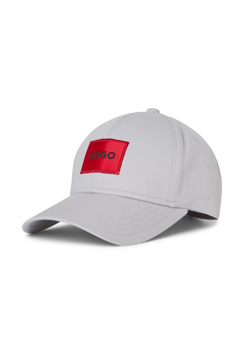 Gorra de béisbol gris con un parche rectangular rojo que lleva la palabra "HUGO" en letras negras. Diseño estructurado con visera curva.