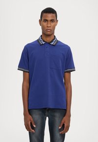 Modrá polo košela so svojbytným telom, krátkymi rukávmi, vzorovaným golierom a jednou náprsnou vreckom. Tkanina je hladká s voľným strihom.