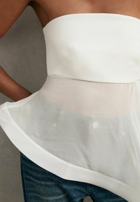 Valkoinen olkaimeton toppi, jossa on läpikuultava beige peplum-yksityiskohta; kangas on sileä ja siinä on hienoinen kiilto, yhdistettynä sinisiin farkkuihin.