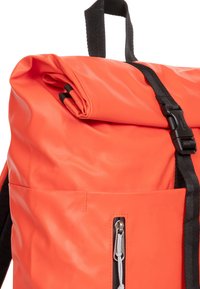 Sac à dos à rabat en orange vif, en nylon lisse. Il dispose d'une sangle noire et d'une boucle réglable, avec une poche zip extérieure.