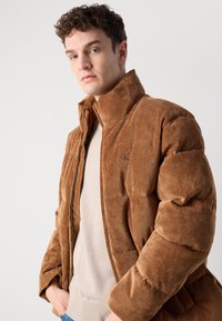 Braune Cord-Pufferjacke mit hohem Kragen, Reißverschluss vorne und subtiler Logodetail auf der Brust. Weicher Stoff und gestepptes Design.