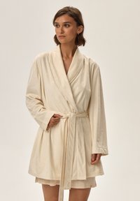 Kremowy szlafrok z kołnierzem typu szal, prostym paskiem w talii oraz teksturowanym wzorem w diamenty, z długimi rękawami i miękkim materiałem.