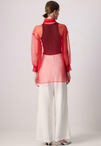 Rote transparente Bluse mit einem einfarbigen roten Rücken, langen Ärmeln und Kragen mit Knopf, kombiniert mit weiten weißen Hosen und silbernen Absätzen.