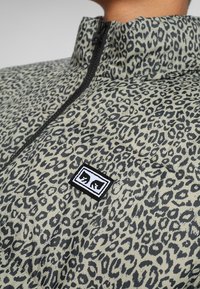 Kurtka puchowa z jasnobeżową bazą i czarnym nadrukiem w cętki lamparta. Posiada wysoki kołnierz i zapięcie na zamek błyskawiczny oraz małą naszywkę z logo na przodzie.