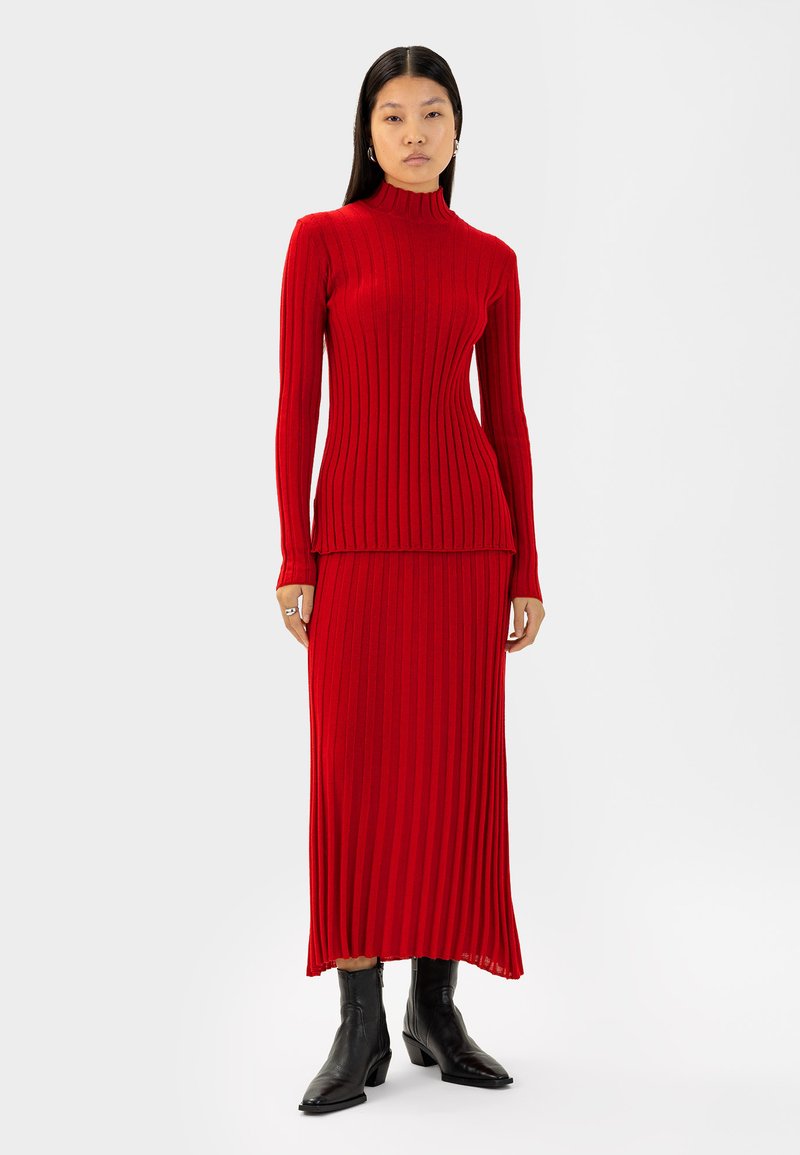 Rotes, geripptes Strickkleid mit langen Ärmeln und hohem Kragen, das eine figurbetonte Oberseite und einen fließenden Rock hat. Kombiniert mit schwarzen Knöchelstiefeln.