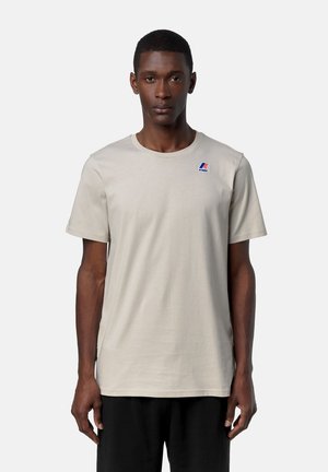 Uomo che indossa una t-shirt beige chiaro a maniche corte con un piccolo logo K-Way blu e rosso sul petto in alto a sinistra, in piedi davanti a uno sfondo semplice.